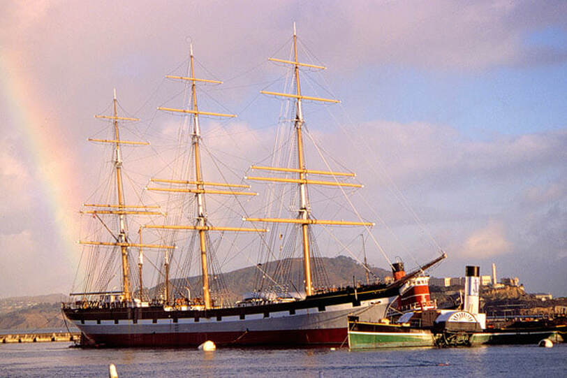 The Balclutha tall ship sits in the harbor with its sails down and a rainbow in the background