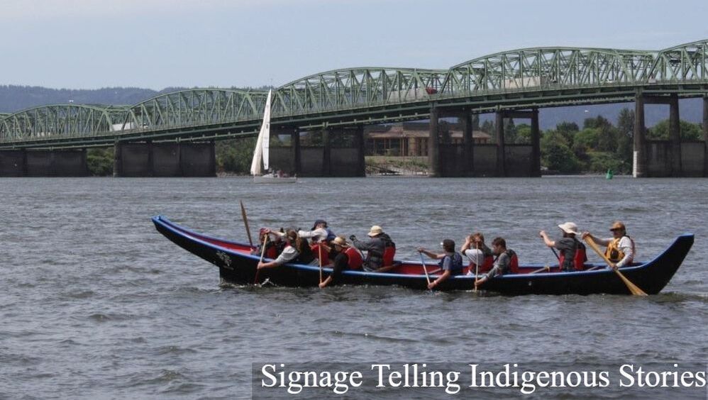 About 10 people sit in a long traditional canoe, across the Columbia River with a bridge in the background