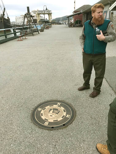 A person in an National Park Service shirt stands next to the installed compass rose with Hyde Street Pier stretching into the background