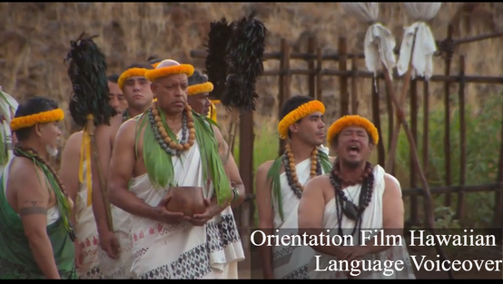 A screenshot from the film Foundation of a Nation; several people wearing traditional togas, scarves, flower headdresses, and large wooden beaded necklaces stand nearby a man who is yelling or singing in ceremony