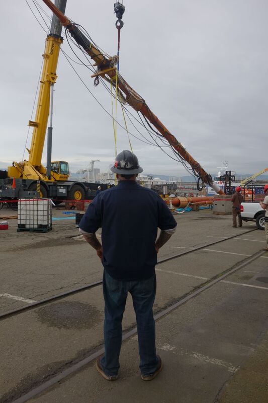 A person with a hard hat faces away from the camera, looking at a large crane lifting a ship mast into place