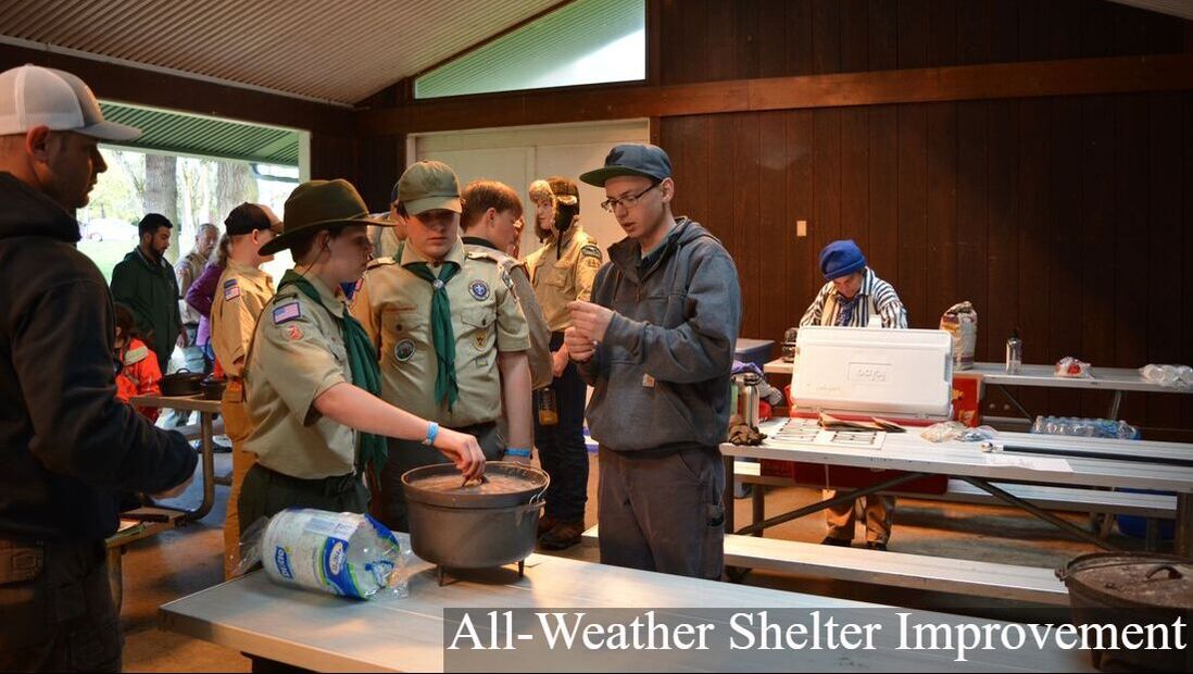 A group of boy scouts in uniform stands around a cast iron dutch oven while other children and adults are seen milling about in the background