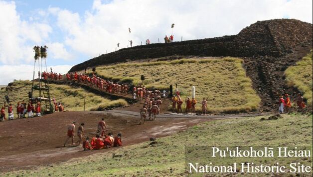 An overview picture of Pu'ukohola Heiau during a ceremony; many people wear traditional Hawaiian dress and are walking up the hill in procession adjacent to a large wooden structure and an ancient edifice made of lava rock