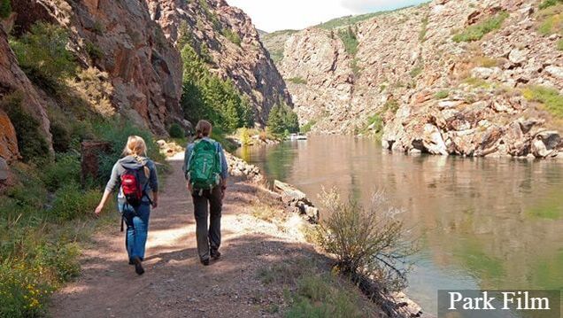 Two hikers walk along a wide trail adjacent to a gentle river with black and red cliffs on either side