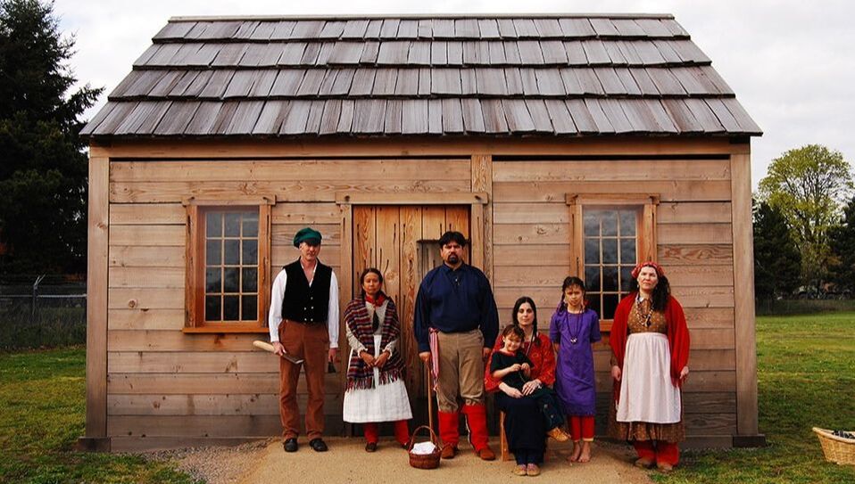 Five people stand in front of a log cabin wearing traditional Indigenous and period attire from the 19th Century 