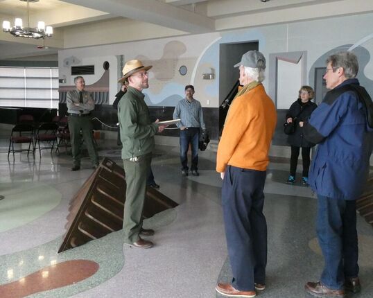 A National Park Service ranger in uniform speaks to a group of visitors while standing amidst murals, sculptures, and decorative flooring in the Aquatic Park Bathhouse