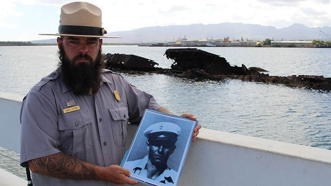 A National Park Service ranger in uniform stands holding a black and white photo of a sailor with the wreckage of the USS Utah half-sunken in the water behind