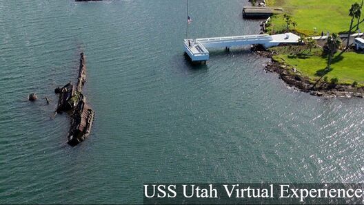 A bird's-eye view of the USS Utah memorial, including a white dock with an American flag and the wreckage of the ship, half sunken in the water
