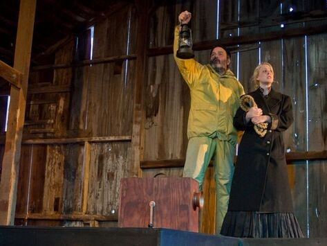 Two actors in costume stand on stage against a wooden barn wall; one is dressed all in yellow and is holding a lantern up, while the other is dressed in a brown dress and clutching a rope