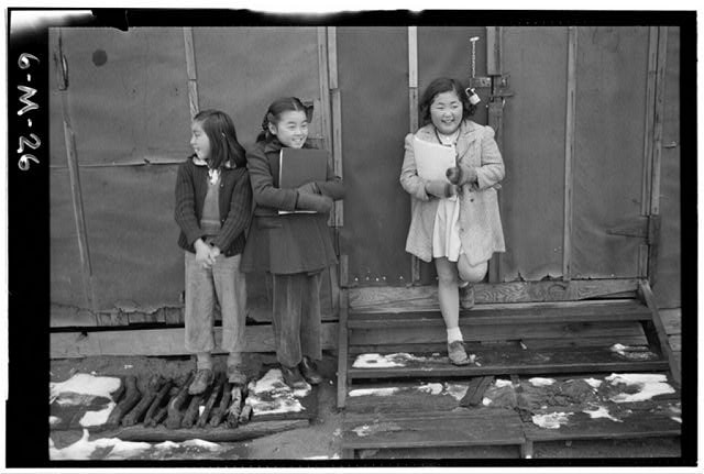 Historic Ansel Adams photo of three children standing in front of a barracks building with school books in hand