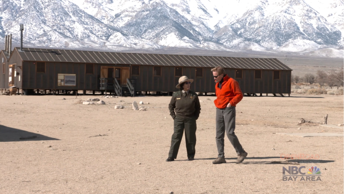 A ranger in uniform walks next to one other person with a barrack-style building and large snowy mountains in the background