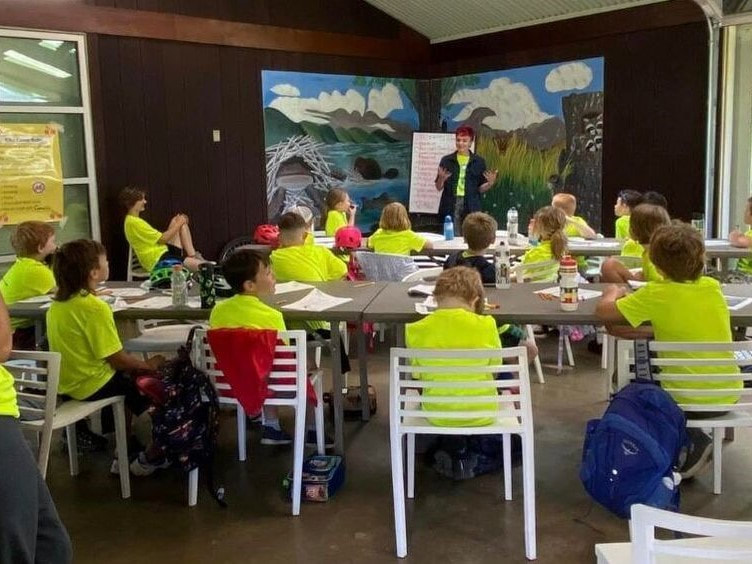 A group of children sit behind tables with papers in front of them facing a speaker with an easel and wildlife mural in the background