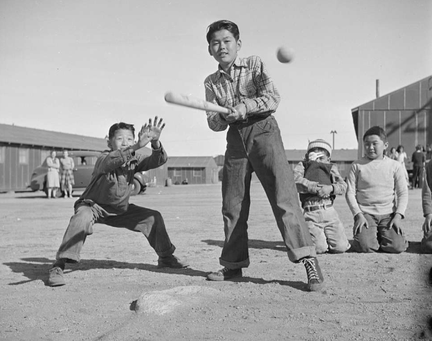 A black and white photo of young children playing baseball with wooden sided barracks in the background