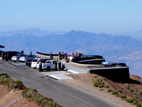 A view from above of Dantes View - a parking lot, a paved overlook with a railing, and an educational sign, all overlooking a valley and mountain range