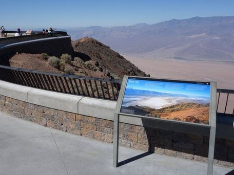 An educational sign sits in front of a stone wall with railing with Badwater Basin and the mountains in the background