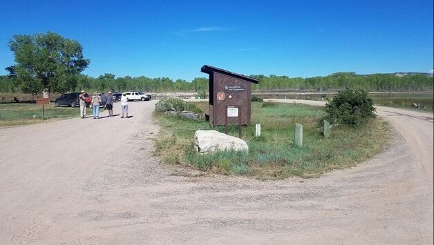 4 visitors walk down a circular roadway with a National Park Service shed and grassy area in the middle. A river is in the background