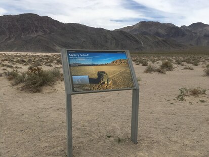 An educational sign with the title "Mystery Solved" and a picture of one of the Racetrack rocks is in front of a desert landscape