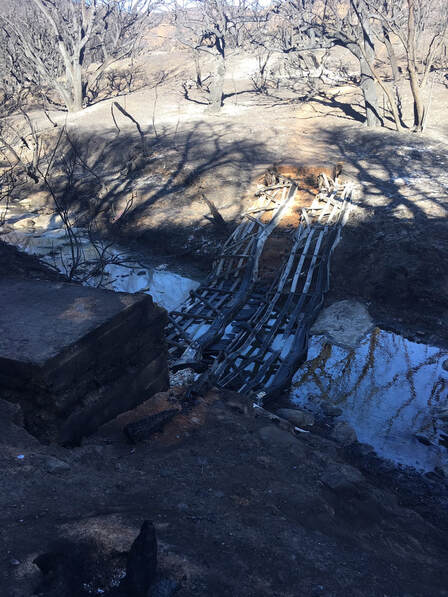 A charred landscape, including the destroyed metal and footings of a bridge draped down into a creek