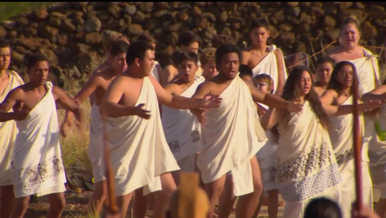 A group of dancers in traditional Hawaiian togas sings with arms outstretched