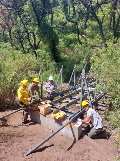 A group of four people wearing hard hats and gloves are adjacent to cement blocks and beams that will hold up a bridge