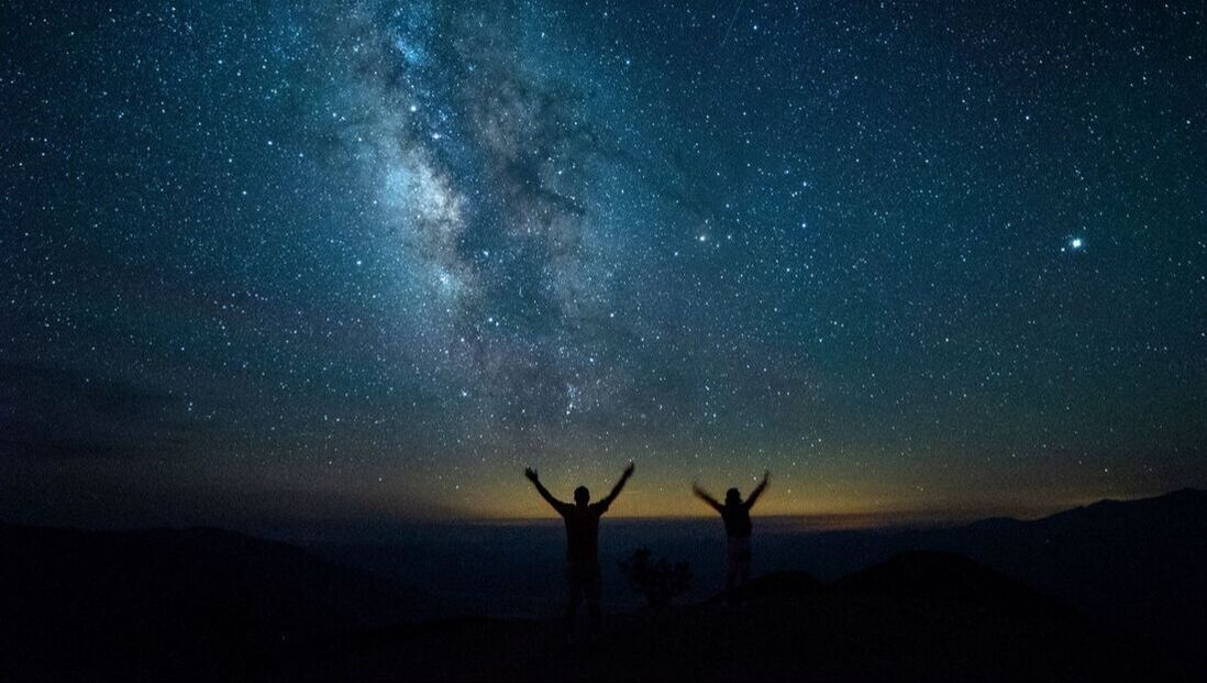 Two people hold up their arms in front of the Milky Way and a starry sky