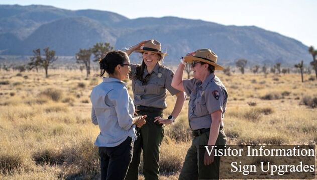 2 National Park Service rangers in uniform hold onto their hats while they talk to a visitor while standing in a joshua tree ecosystem