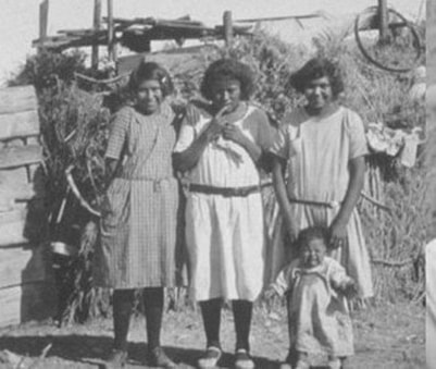 A black and white photograph of 3 adults and one child standing outside a structure made of vegetation
