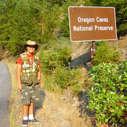 A young person with a vest and hat full of junior ranger badges stands in front of some bushes and an Oregon Caves National Preserve sign