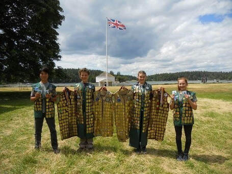 A group of four children stand in a line facing the camera, each wearing and holding vests covered in Junior Ranger badges; a meadow, fort-like structure, and the British flag are in the background