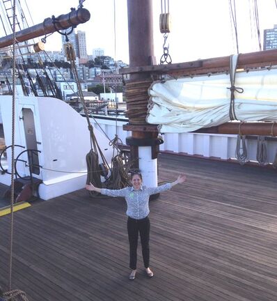 Person stands with arms in the air on wooden deck of ship below large mast with sail in closed position