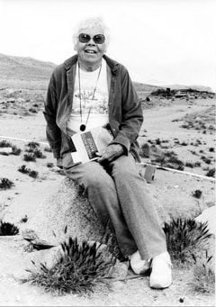 Black and white photo of Mary Dedecker, who sits on a rock with a book in her hand with a desert landscape behind