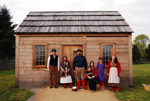Several people are in front of a wooden cabin wearing attire from the 19th century, as well as Indigenous adornments