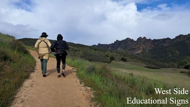 Two people walk along a wide dirt path set amidst a grassy meadow with the Pinnacles rock formation in the background