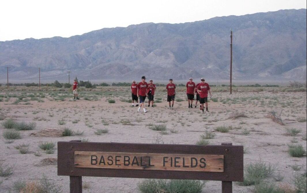 A group of young people in red and black baseball uniforms walks across an empty lot with a wooden sign in the foreground that says 