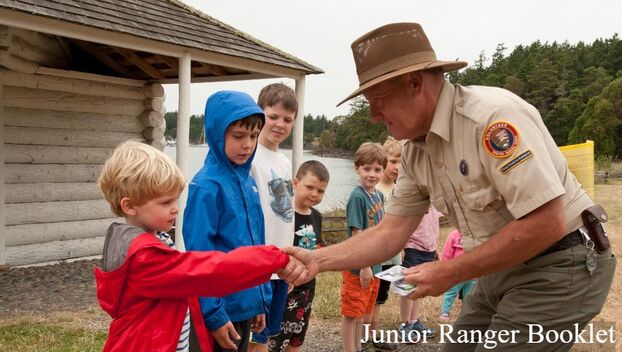 A person in a National Park Service volunteer uniform shakes hands with a child standing in a line among other children with a cabin, lake, and trees in the background