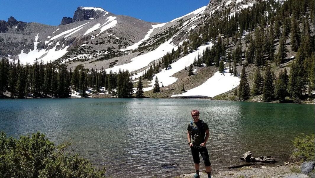 Person stands in front of Stella Lake with a mountain ridge of snow and rock behind