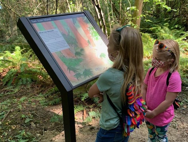 Two young visitors wearing masks read a wayside educational sign titled, 