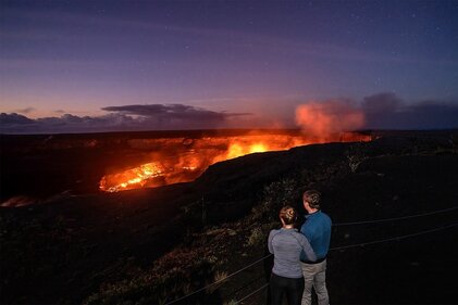 Two people stand arm in arm watching red glowing lava and steam coming off a dark volcanic landscape