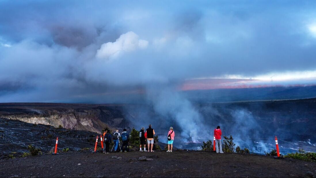 A group of visitors stands behind a coned line looking at smoke coming out of a crater