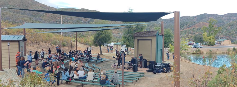 An amphitheater covered in shade is filled with people watching a band play on a small stage with the edge of the lake and rolling hills in the background