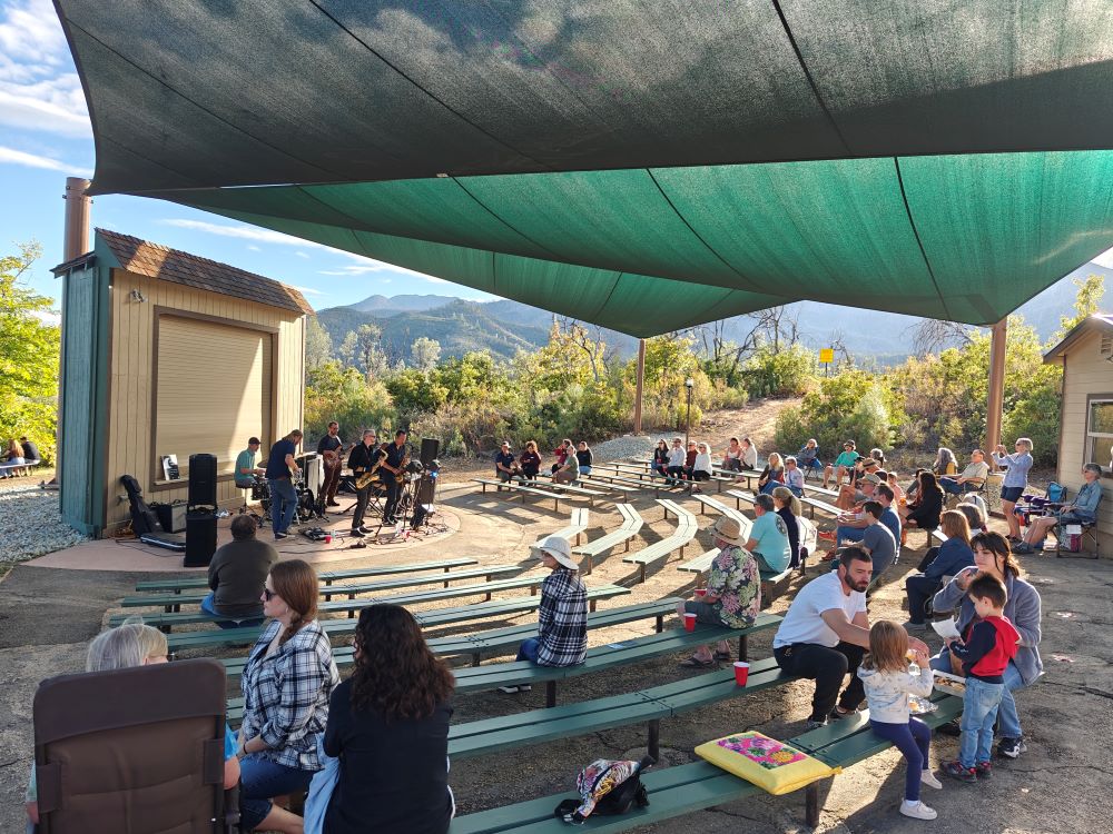 A band plays on the Whiskeytown amphitheater stage with about 50 people in the benches watching the show under large shade canopies