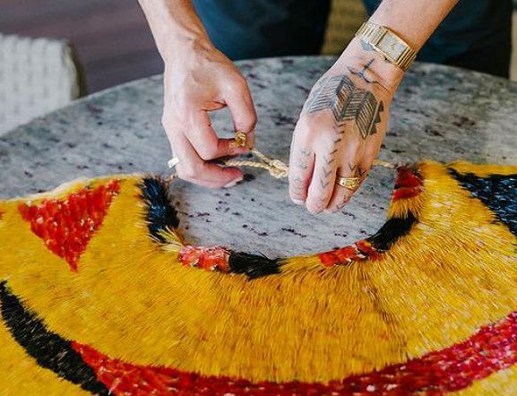 Two hands are shown tying a knot of twine connected to a colorful garment made with feathers