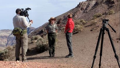 A person films a ranger talking to another person in a rocky desert landscape