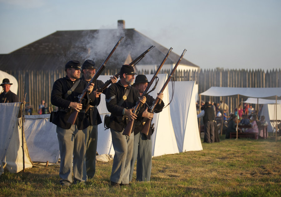 4 people wearing historic military uniforms hold rifles with smoke coming off of them inside of a fort with tents in the background