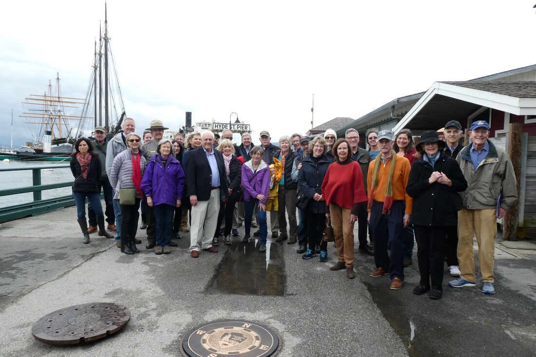 A group of about 30 people stands behind the installed compass rose with Hyde Street Pier and historic ships in the background.