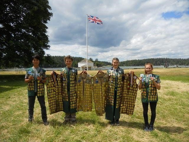 Four children stand astride one another holding and wearing vests with Junior Ranger badges with a fort-like building and British flag behind them