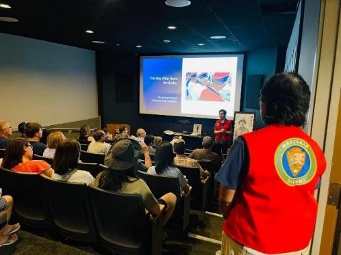 A group of people watches a presentation in an auditorium; a volunteer with the NPS logo on the back of their vest stands in the foreground watching the presentation