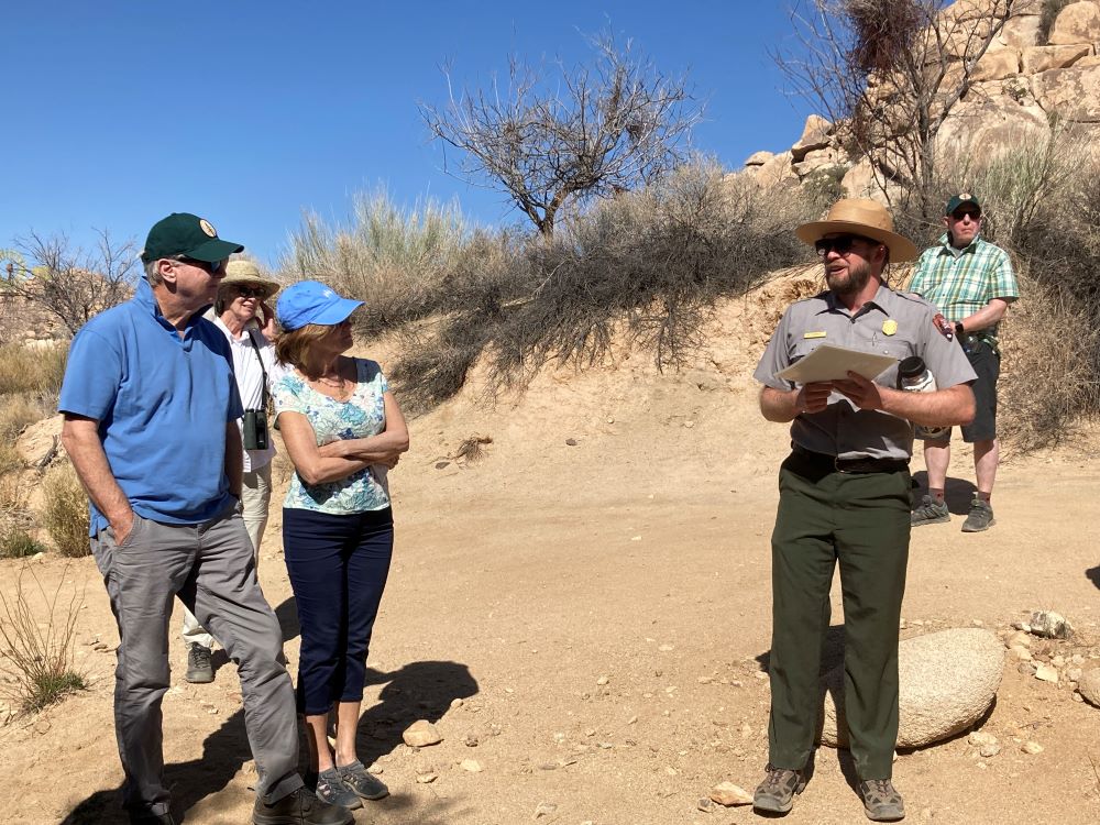 Two people stand looking at a National Park Service ranger in uniform giving a talk with a desert landscape in the background
