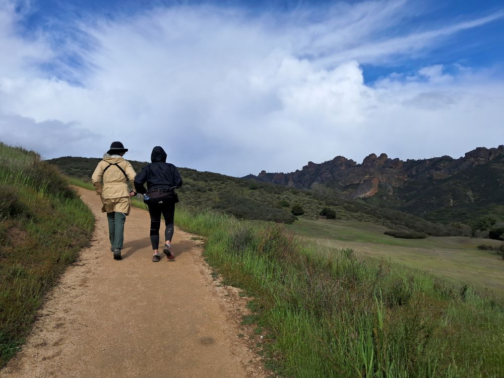 Two people walk along a wide dirt trail amidst green grassy hills with rocky pinnacles in the background