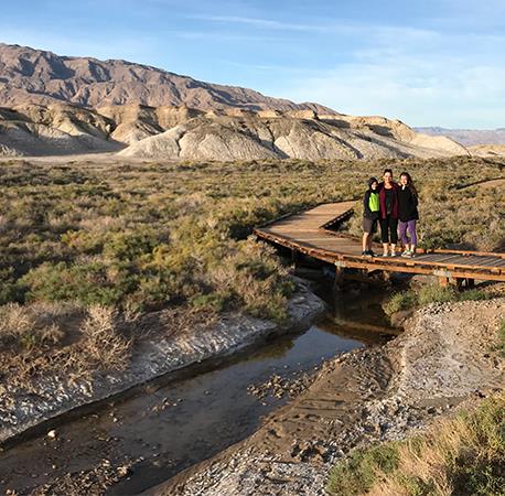 Three visitors stand on the Salt Creek Boardwalk with the narrow band of creek below them and a desert landscape behind